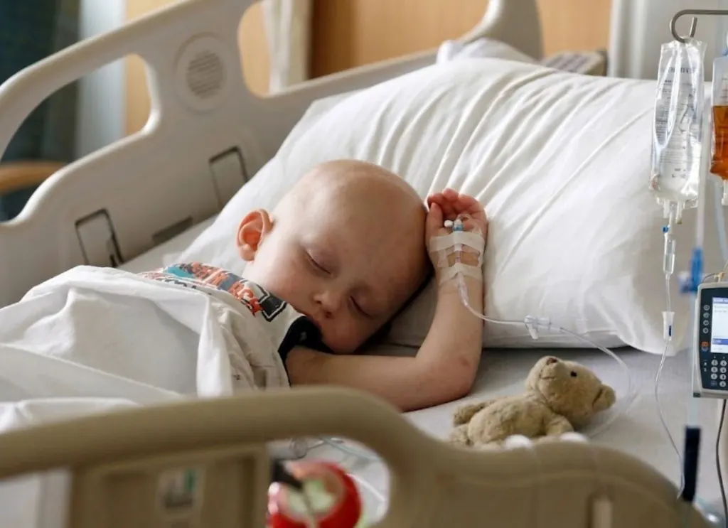 Kind liegt in einem Krankenhausbett mit Infusion und Kuscheltier.